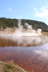 Photo of a hot spring with a reddish brown lake bottom in the foreground and smoke on the water in the background